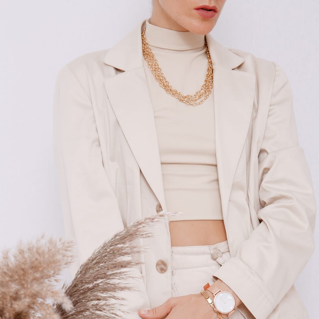 A woman dressed in a stylish, neutral-toned casual outfit. She wears a cream blazer over a matching crop top, paired with cream pants. Accessories include layered gold necklaces, a gold watch on her wrist, and a hint of makeup with soft pink lips.