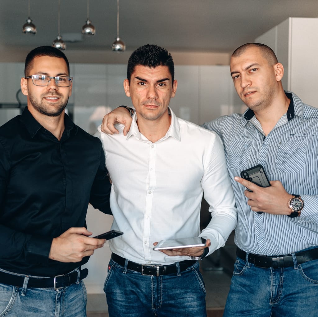 Three men standing together indoors, dressed in casual button-down shirts, with two holding smartphones and one resting his hand on another's shoulder, exemplifying a stylish and confident look.