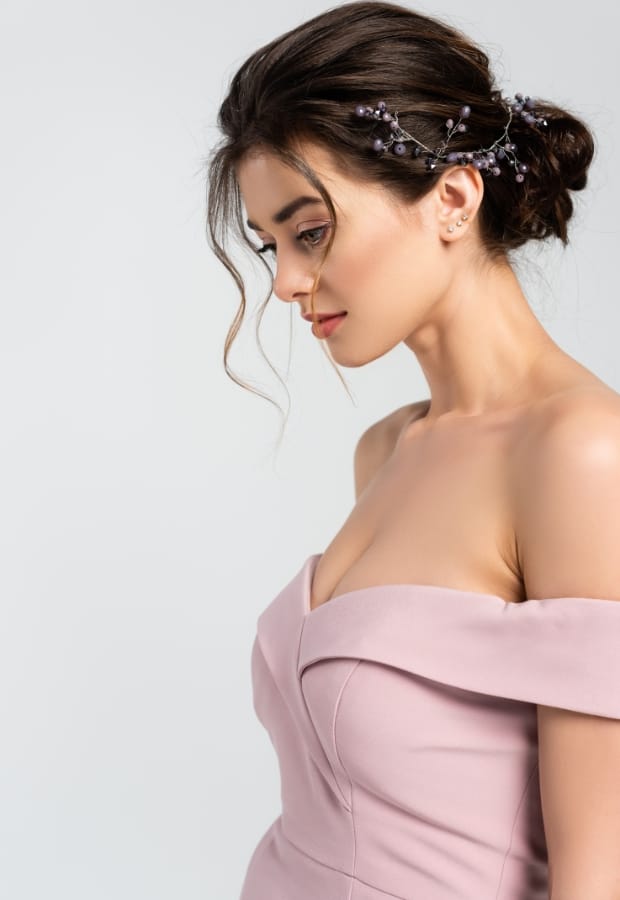 A young woman with styled brown hair adorned with delicate purple floral hair accessories, wearing a strapless blush pink prom dress, looking down softly with a neutral background.