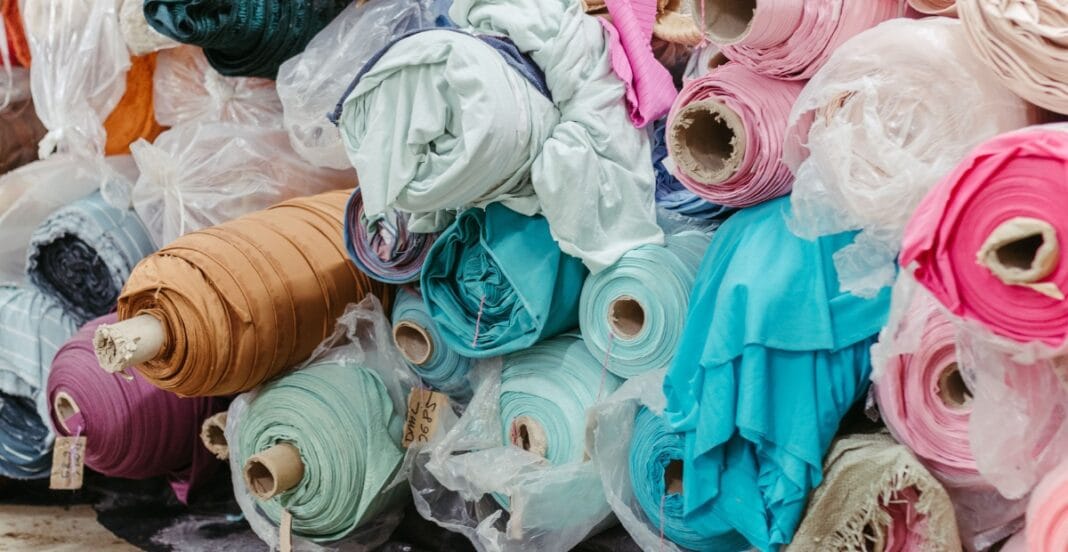 A collection of colorful fabric rolls and clothing materials, including pink, blue, green, black, and beige, piled together and wrapped in plastic.