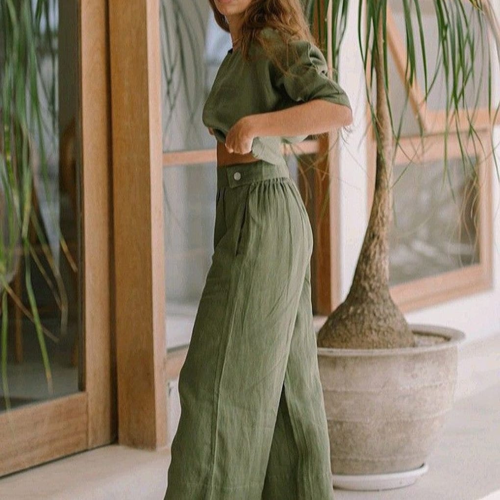 Woman wearing a matching olive-green crop top and wide-leg trousers, standing indoors next to a large potted plant and wooden-framed glass doors.