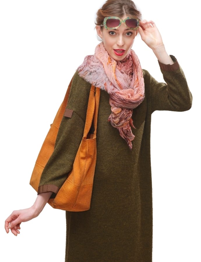 A woman wearing a long, dark green coat with a textured cotton fabric, accessorized with sunglasses, a pink patterned scarf, and a large orange tote bag.