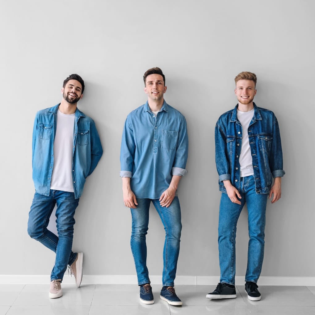 Three men standing against a plain light grey wall, dressed casually in denim and chambray shirts and jeans, showcasing modern men's fashion trends.