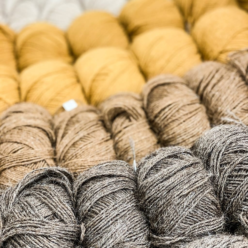 Close-up image of multiple balls of yarn arranged in rows, with shades of yellow, light brown, and dark brown.