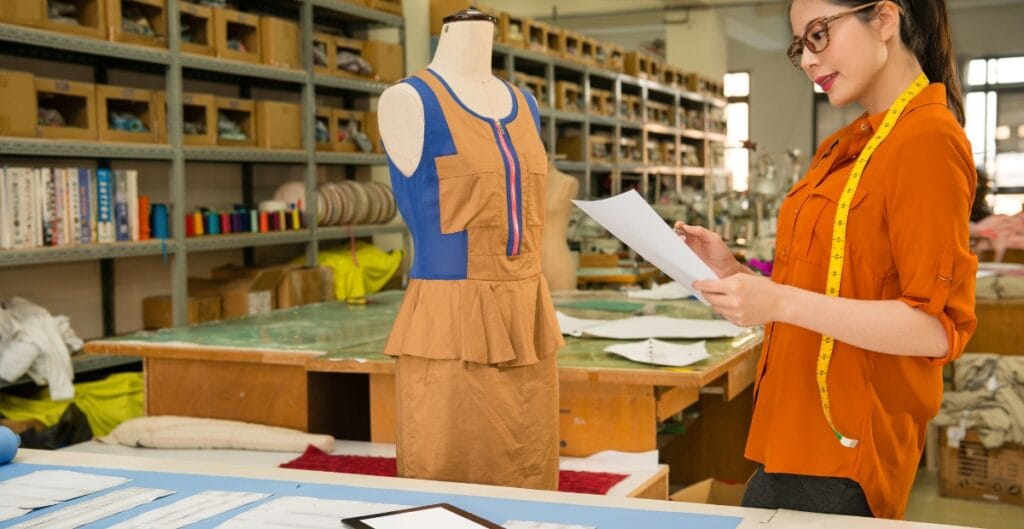 A woman in an orange shirt with glasses and a measuring tape around her neck is working in a textile or fashion design studio, reviewing papers next to a dress form fitted with a colorful garment, with shelves filled with fabrics and sewing supplies in the background.