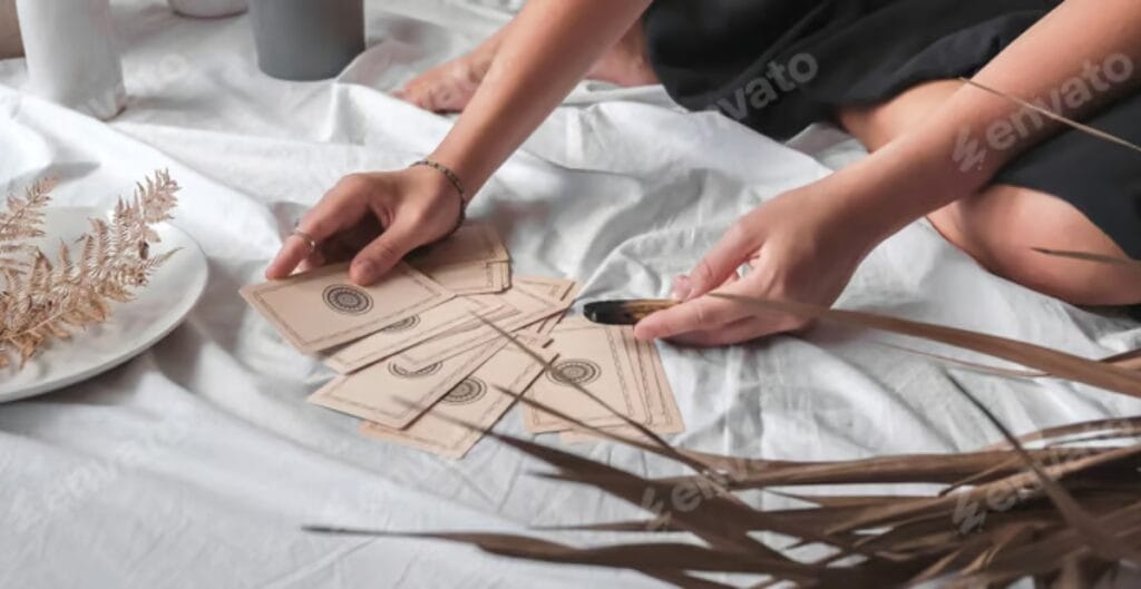 Two hands are seen arranging tarot cards on a white fabric surface. One person is holding a fan of tarot cards, while the other is holding a single card. There are dried plants nearby, and a white plate with more dried foliage on it.