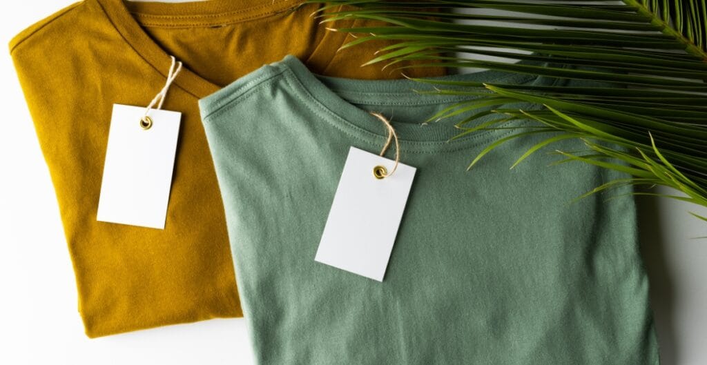 Two folded sweatshirts, one mustard yellow and one muted green, with blank white tags attached, partially covered by green palm leaves.