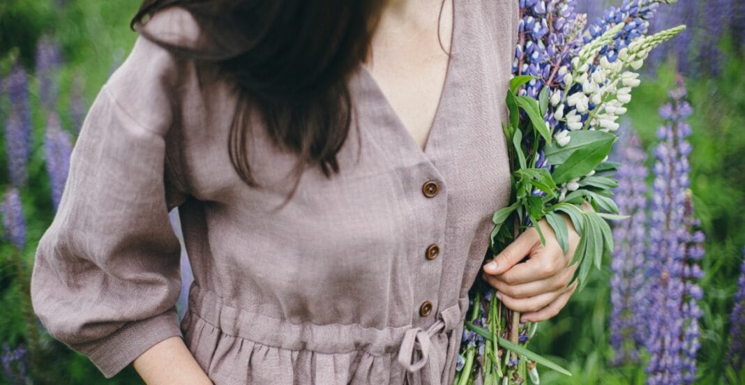 A woman holding a bouquet of purple and white flowers in a field of tall purple flowers, wearing a light brown dress with buttons.