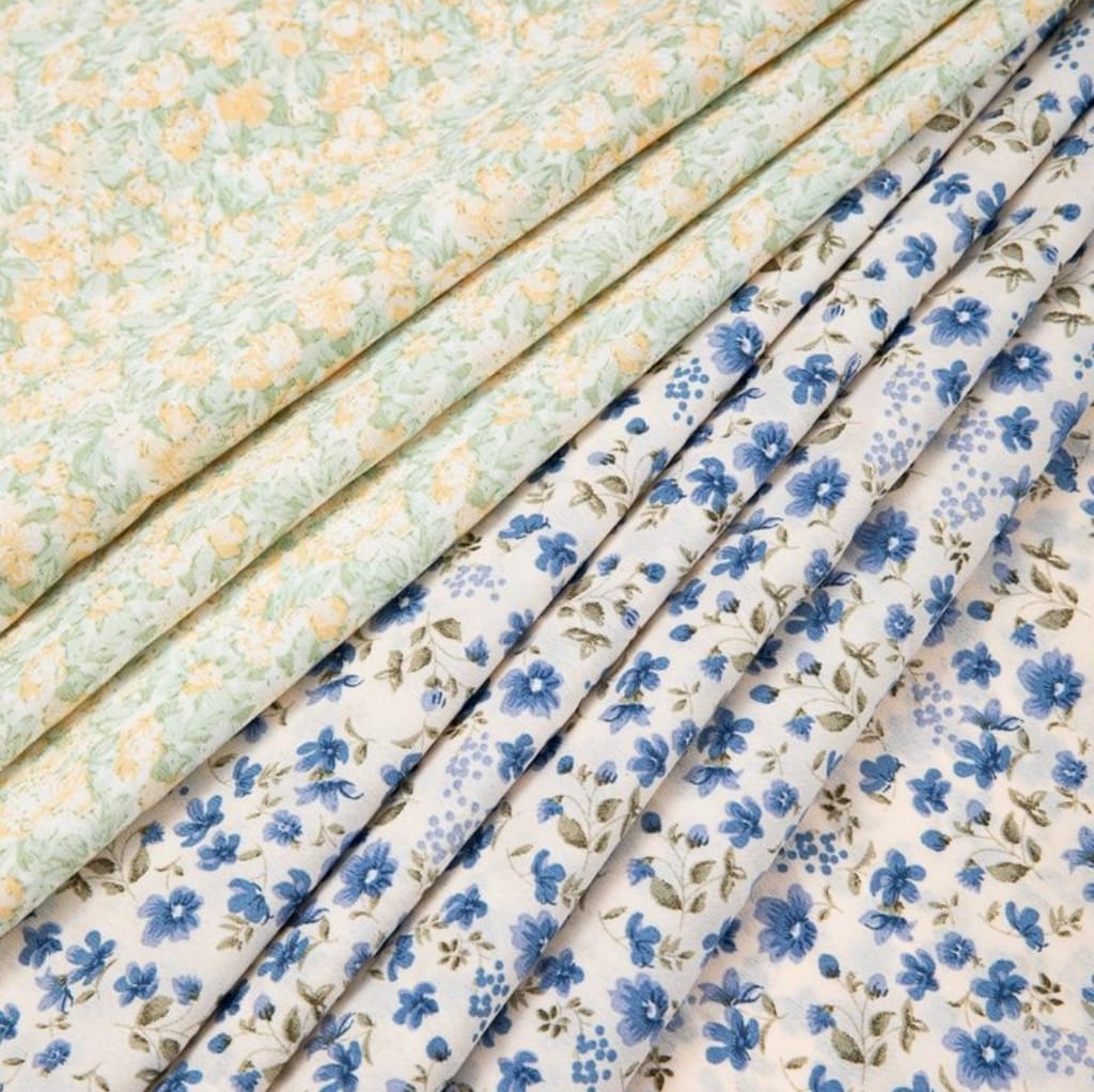 Close-up image of several folded fabrics featuring floral cotton prints in pastel yellow, green, and blue colors with delicate, intricate flower patterns.