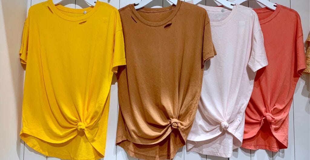 Four t-shirts hanging on a rack, each tied in a knot at the front, in yellow, brown, white, and red colors.