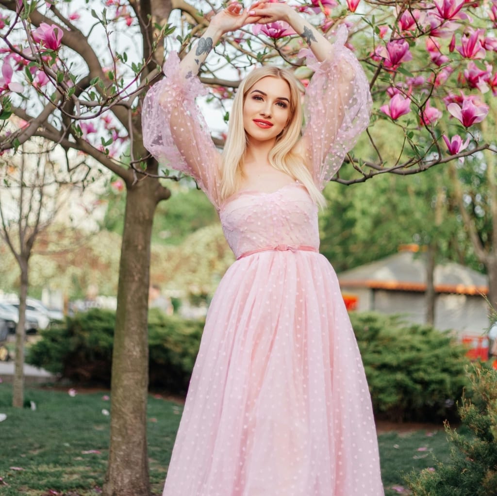 A woman standing outdoors under a blooming tree, wearing a flowing pink dress with sheer puffed sleeves, surrounded by vibrant pink blossoms.