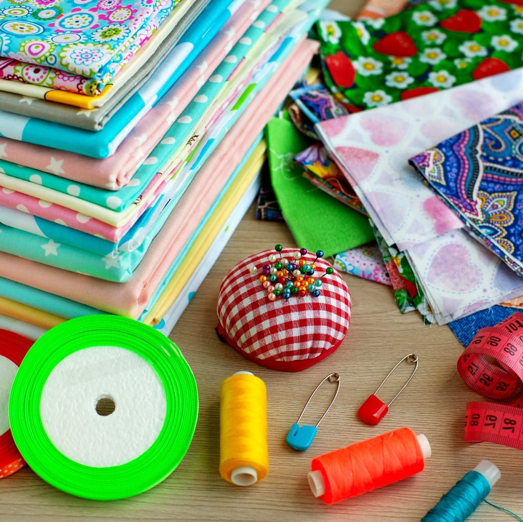 A collection of colorful cotton fabrics with playful patterns, sewing pins on a red and white checked pincushion, various spools of thread in yellow, orange, and turquoise, measuring tape, safety pins, and green elastic on a wooden surface, indicating a sewing project setup.