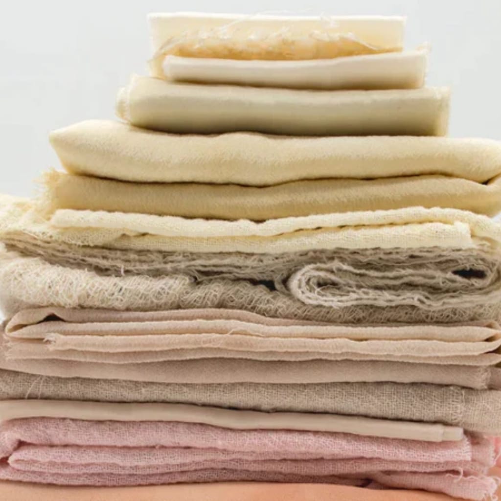 A neatly stacked pile of various folded fabrics, mostly in neutral or pastel tones, with some textured and smooth materials, arranged in layers from top to bottom.