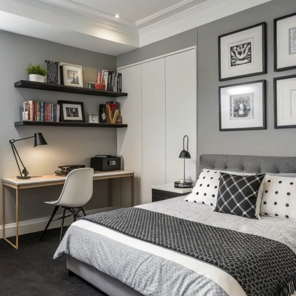 : A stylish bedroom featuring a neatly made bed with black, white, and gray patterned pillows, a gray upholstered headboard, a modern white bedside table with a black lamp, and a white desk with a white chair. Two black floating shelves hold books, framed photos, and decorative items, set against soft gray walls with a gallery of black-and-white framed artwork.