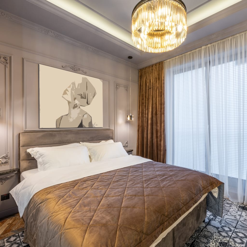 A luxurious bedroom featuring a beige upholstered headboard, white bedding, a brown quilted blanket, framed abstract artwork above the bed, elegant wall lighting, and floor-to-ceiling windows with white sheer curtains and gold drapes. A modern, golden chandelier hangs from the ceiling, adding warmth and sophistication to the space.