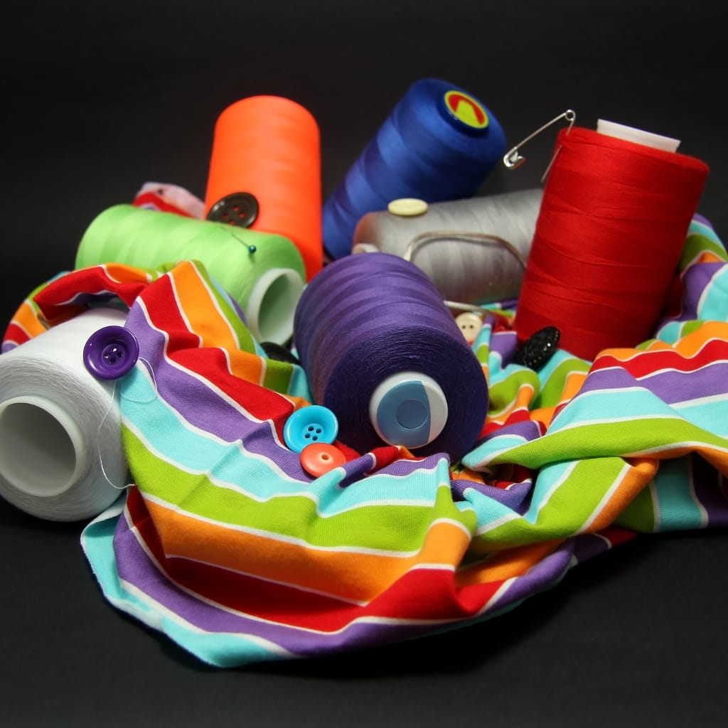 A colorful, striped, lightweight fabric with a shiny, smooth surface, surrounded by spools of bright colored thread and buttons.