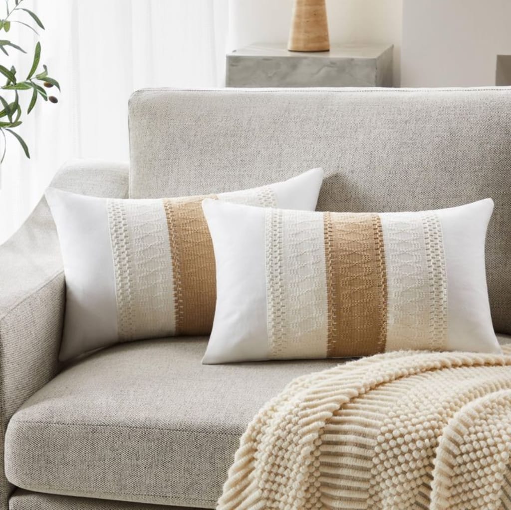 Two decorative pillows with beige and white woven patterns rest on a light-colored sofa, with a cozy textured blanket draped over the armrest and a modern, minimalist interior in the background.
