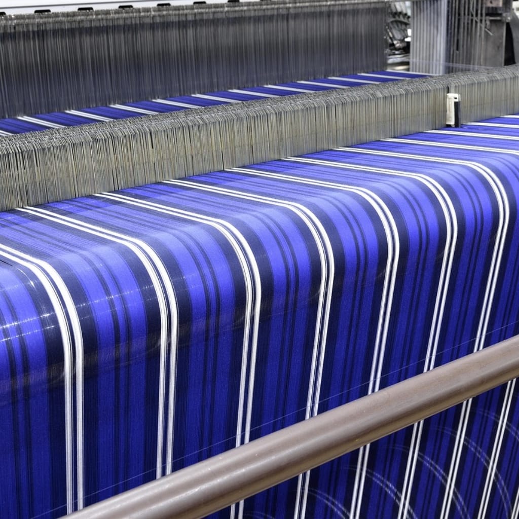 Close-up image of an industrial weaving machine producing vibrant blue fabric with white and black stripes. The fabric is being woven on large rollers, with the machine's components visible at the top.