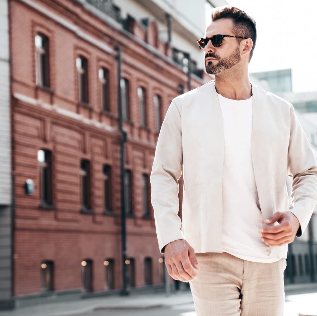 A stylish man with sunglasses walking outdoors in a modern urban area, dressed in a light-colored jacket and beige pants.