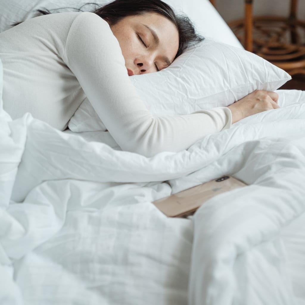 A woman is peacefully asleep on a bed with white bedding, resting her head on a pillow, with a smartphone lying on the bed next to her.