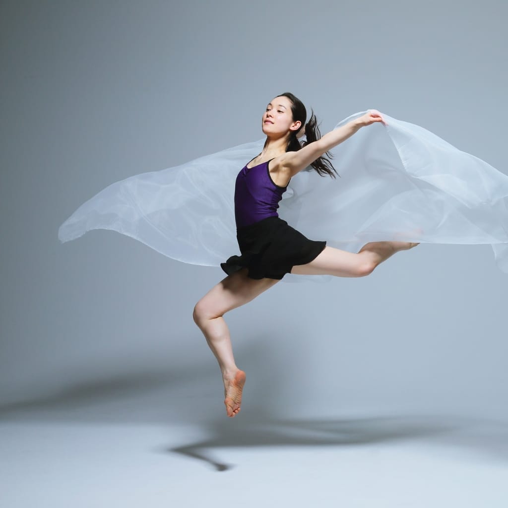 A female dancer mid-air, with arms outstretched and a flowing white fabric behind her, against a plain gray background.