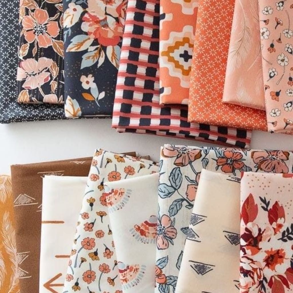 A selection of various cotton print fabrics, each with different floral and geometric patterns in shades of peach, pink, navy blue, white, and brown, neatly folded and arranged on a white surface.