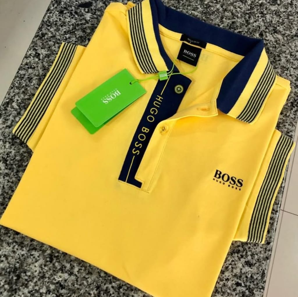 A bright yellow Hugo Boss polo shirt with navy accents, neatly folded on a granite countertop. The shirt features a navy collar with yellow and navy striped detailing, a button placket with "HUGO BOSS" embroidered in yellow on the navy strip, and the "BOSS" logo on the chest.