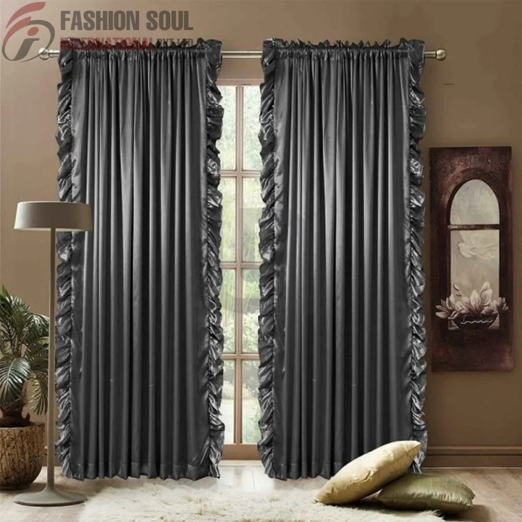 A pair of dark gray, shiny curtains with ruffled edges hanging in front of a window. There is a beige wall with a decorative framed wall art piece on the right, a standing lamp on the left, and a potted plant next to the lamp. Two cushions, one gold and one green, are placed on a white rug on the floor.