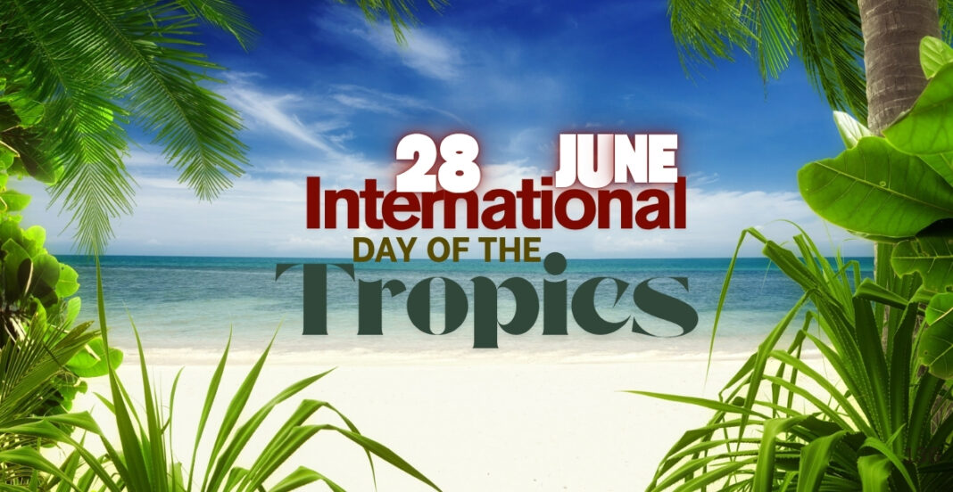 A tropical beach scene with green palm and broadleaf plants framing a sandy shore and ocean under a partly cloudy sky, with overlaid text celebrating June 28th as International Day of the Tropics.