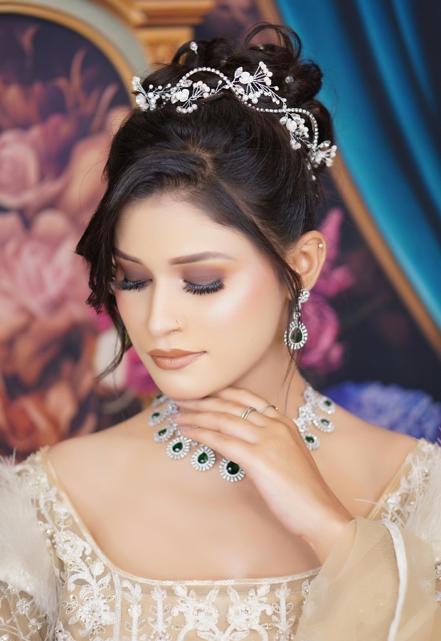 Close-up of a woman adorned with intricate jewelry, including a pearl and green gemstone necklace, matching earrings, and a decorative tiara, all complementing her elegant attire.