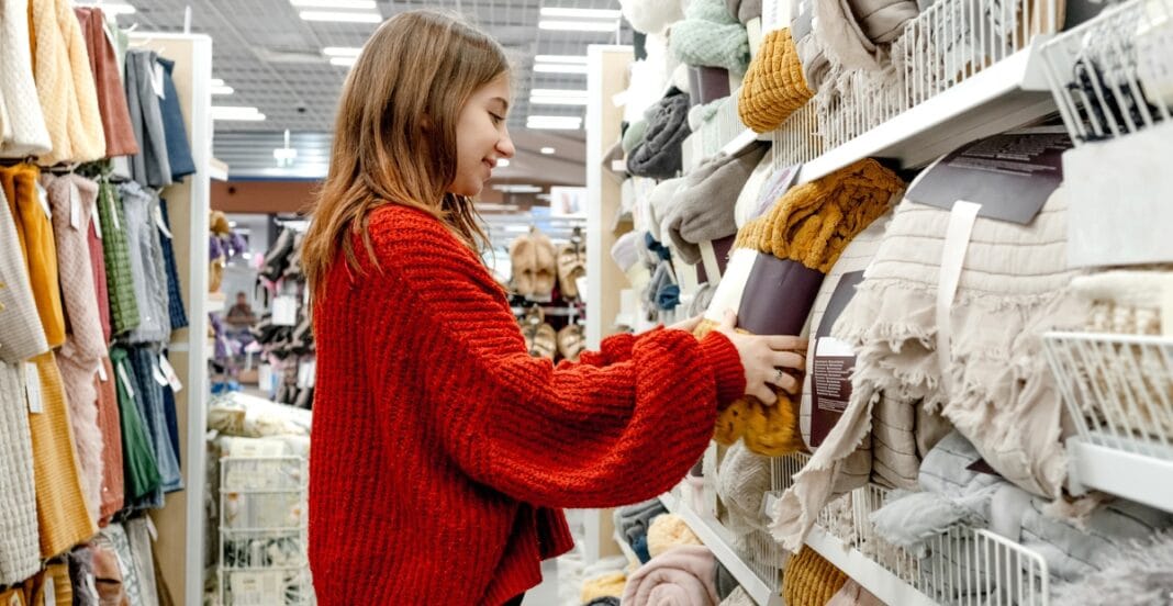 A young woman in a red sweater shopping for textiles or blankets in a store aisle filled with folded fabric and home textiles.