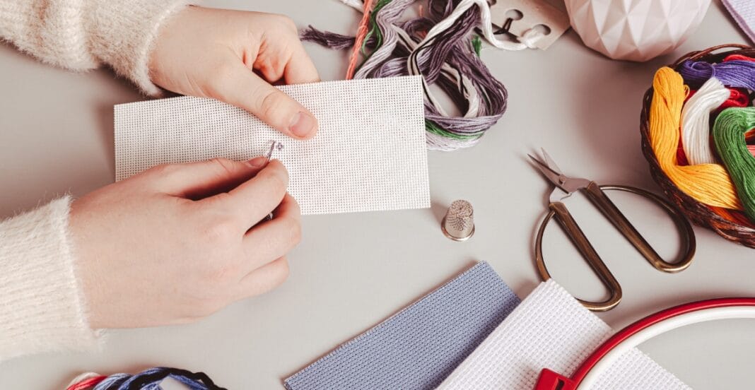 A person stitching miniature canvas on a table surrounded by cross-stitch supplies, including colorful embroidery threads, scissors, a thimble, and fabric.