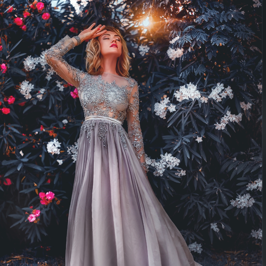 A woman in an elegant gown with a sheer, intricate lace top and a flowing satin skirt, standing amidst lush greenery and blooming flowers with sunlight filtering through.