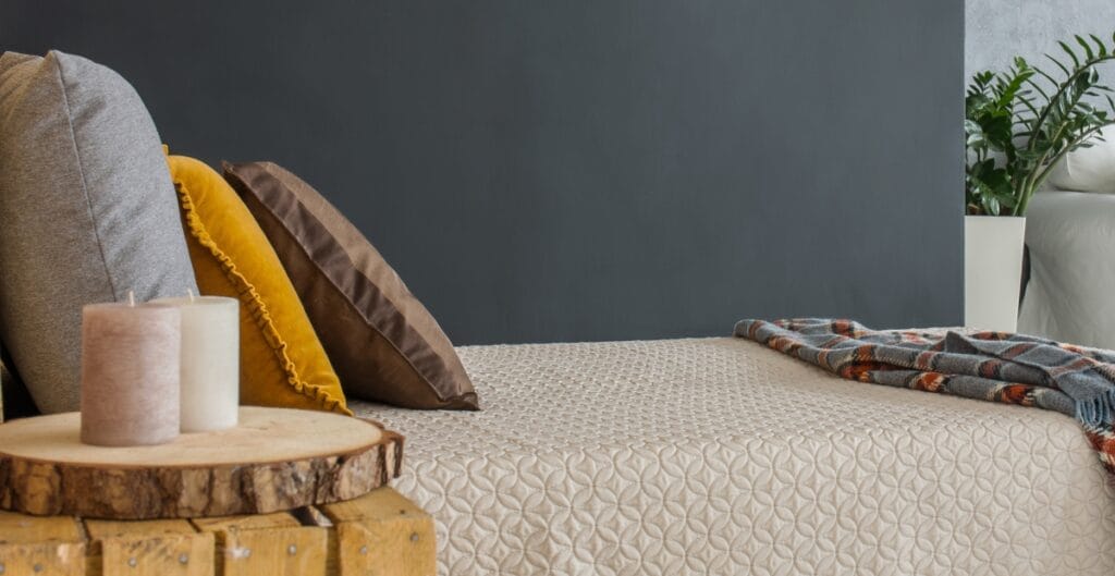 A cozy seating area with a beige textured fabric surface, layered with three throw pillows in gray, yellow, and brown. There is a small round wooden table holding three candles. To the right, part of a plaid blanket is draped over the seating, and in the background, a dark gray wall and some green plants in white pots are visible.