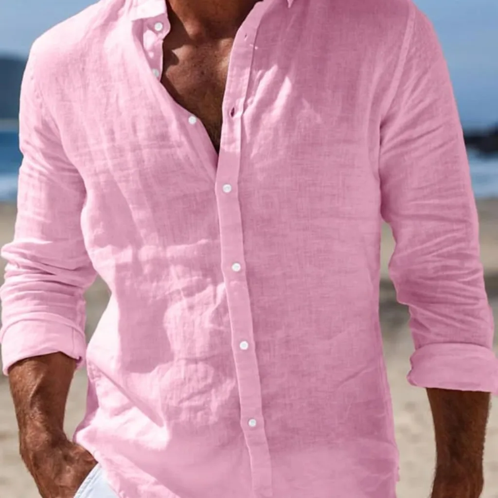 A close-up of a man wearing a light pink linen shirt with the top few buttons undone, standing on a beach with sand and ocean in the background. The sleeves are rolled up to the elbows.