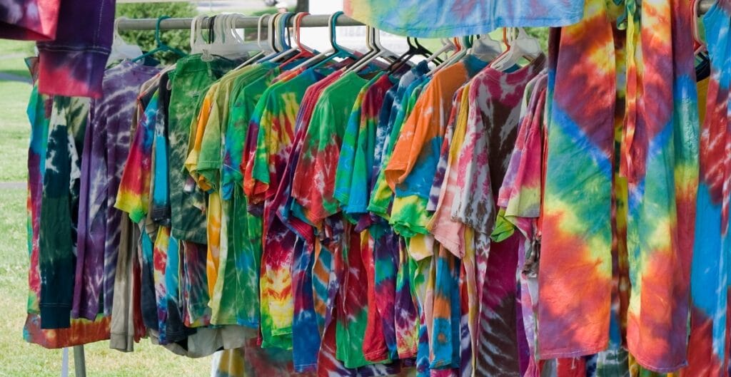 Colorful tie-dye shirts hanging on a clothes rack outdoors with a grassy background.