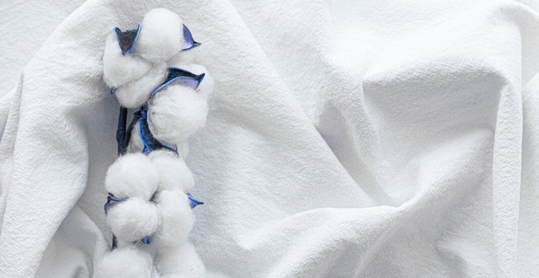 A close-up photograph of long-stapled cotton fabric with a soft, textured surface and a white, slightly crinkled appearance. Cotton bolls are placed on the fabric, highlighting the natural origin of the material.