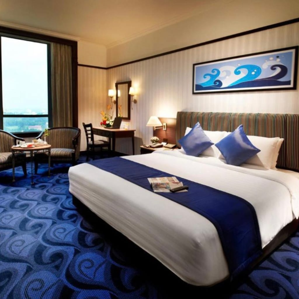 A cozy hotel room with a large bed, striped headboard, two blue pillows, a wall-mounted artwork of stylized ocean waves, a seating area with chairs and a small table, a dressing table, and a large window with curtains, in a well-lit setting.