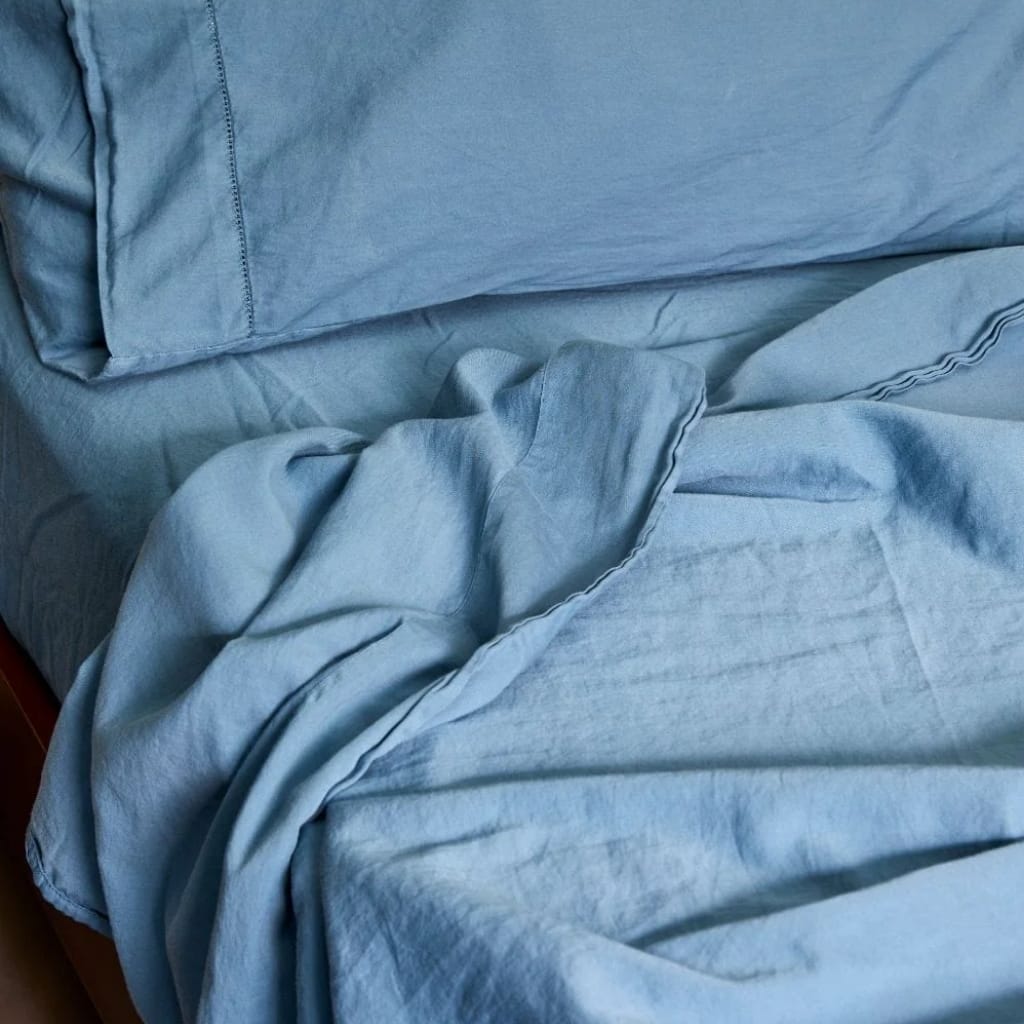 A neatly made bed with light blue bed sheets, pillowcases, and a blanket. Some of the sheets are slightly wrinkled, and part of the bedding is folded or crumpled on the right side.