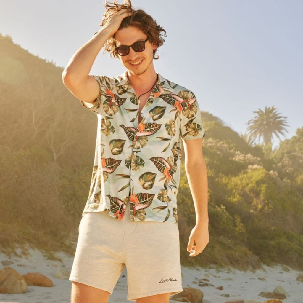 A young man with tousled hair, wearing sunglasses, a colorful floral beach shirt, and beige swim trunks, smiling and running a hand through his hair on the beach.