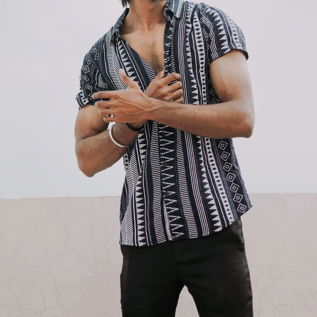 A man wearing a stylish, short-sleeved summer shirt with a black and white geometric tribal pattern, paired with black pants, standing against a plain light background.