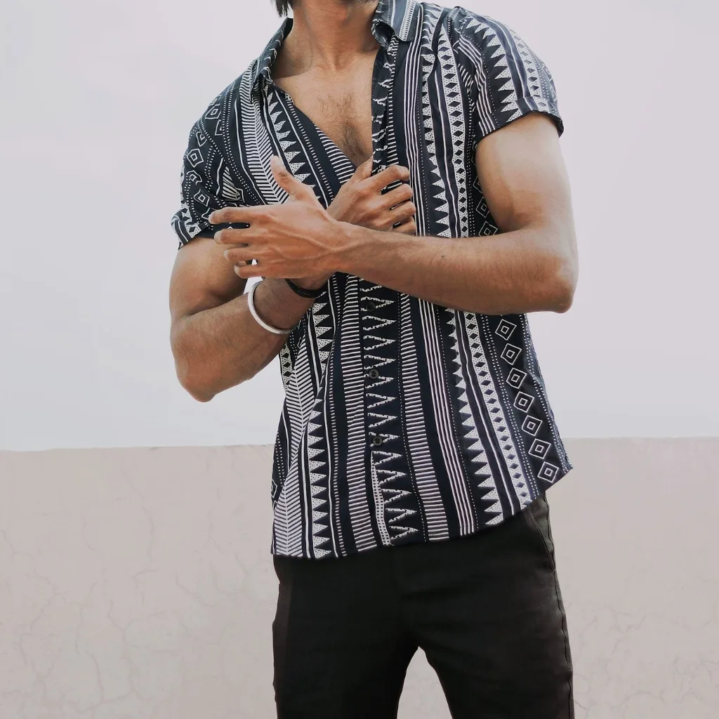 A man wearing a stylish, short-sleeved summer shirt with a black and white geometric tribal pattern, paired with black pants, standing against a plain light background.