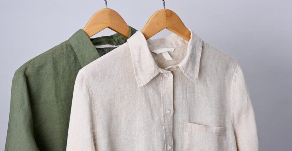 Two men's linen shirts, one green and one white, hanging on wooden hangers against a plain grey background.