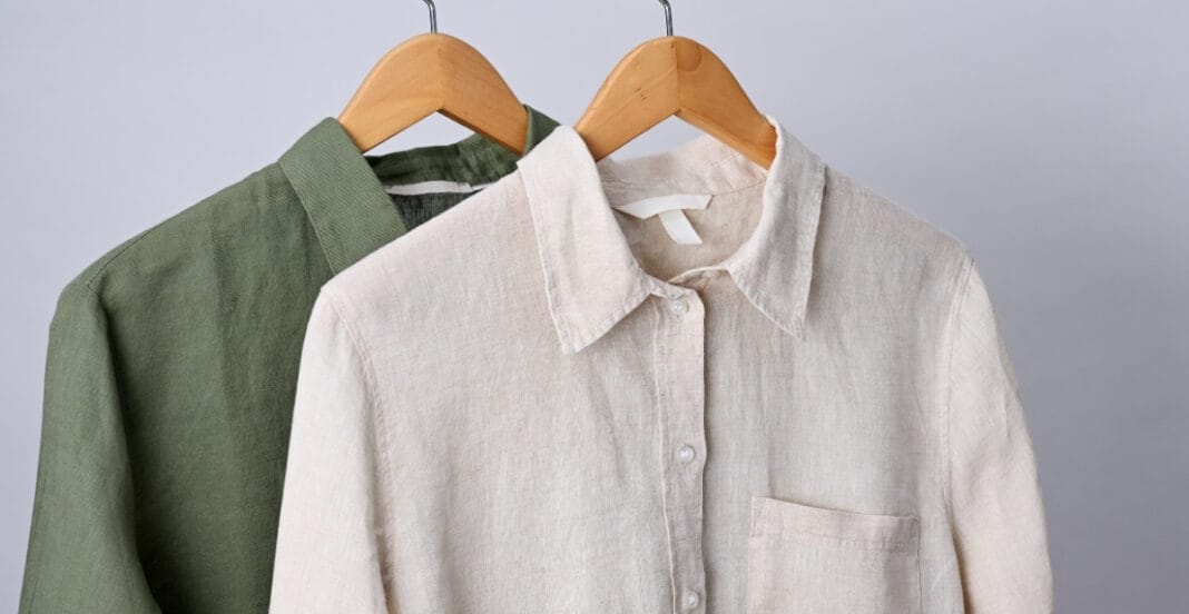 Two men's linen shirts, one green and one white, hanging on wooden hangers against a plain grey background.