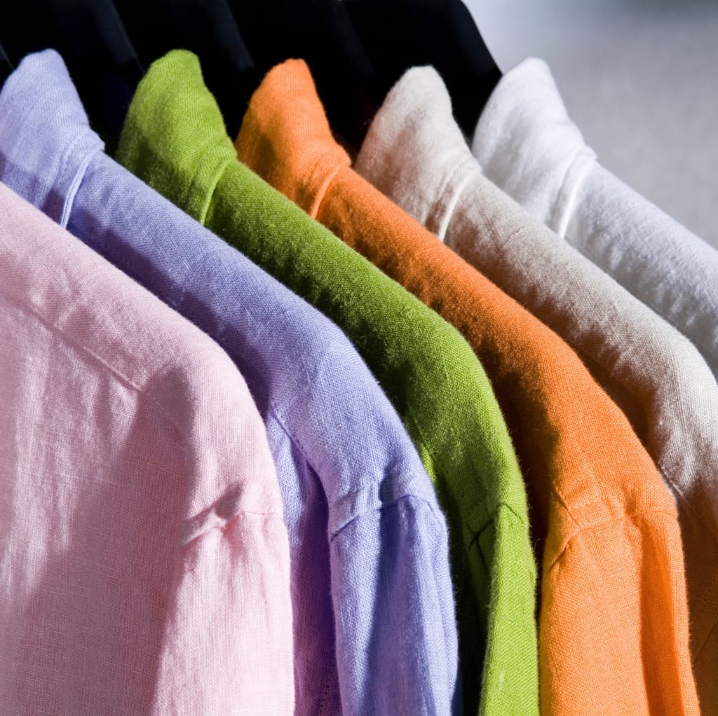 A row of men's linen shirts in pastel and neutral colors hanging on a rack, including pink, lavender, green, orange, beige, and white.