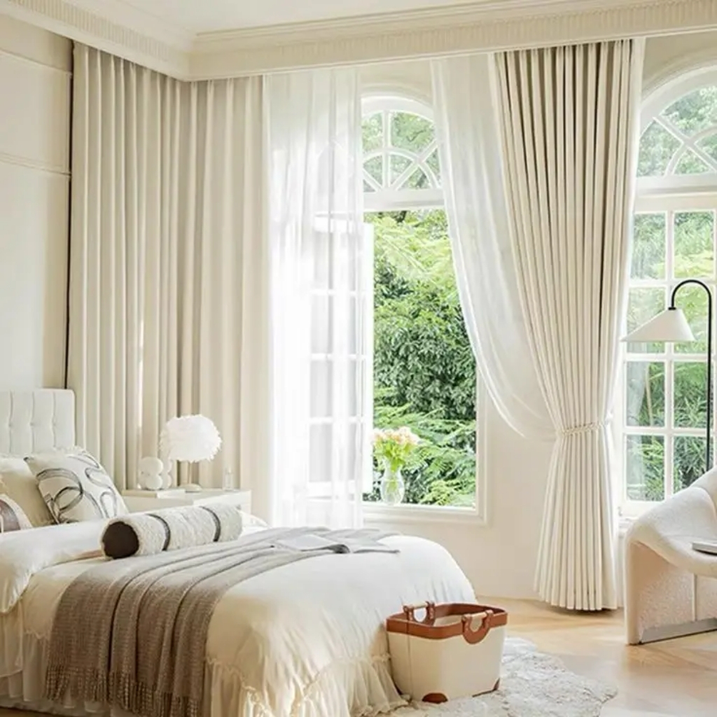 A bright, elegant bedroom featuring large arched windows with white curtains and sheer drapes, overlooking lush greenery outside. The bed has white bedding with a striped bolster pillow and a cozy throw blanket. Near the window is a white armchair, and a small beige suitcase sits on the light wooden floor.