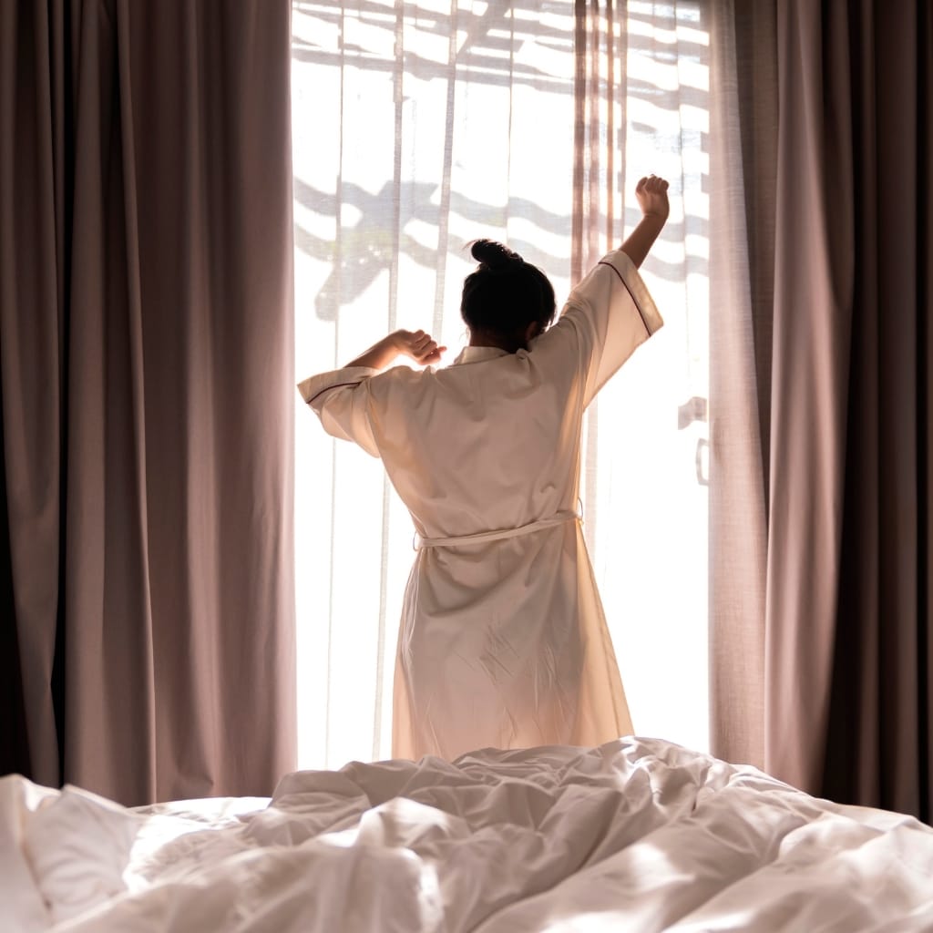 A person standing by a window with sheer curtains, stretching with arms raised, seen from behind. They are wearing a light-colored robe, and sunlight filters through the curtains, creating shadows of tree branches on the window.
