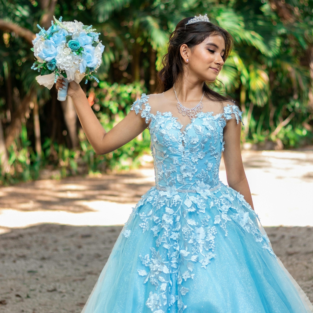 A young woman in a beautiful light blue ball gown with intricate floral lace details, holding a matching bouquet of blue and white flowers, standing outdoors amidst lush greenery. She wears a sparkling tiara and matching jewelry, with a graceful and confident expression.