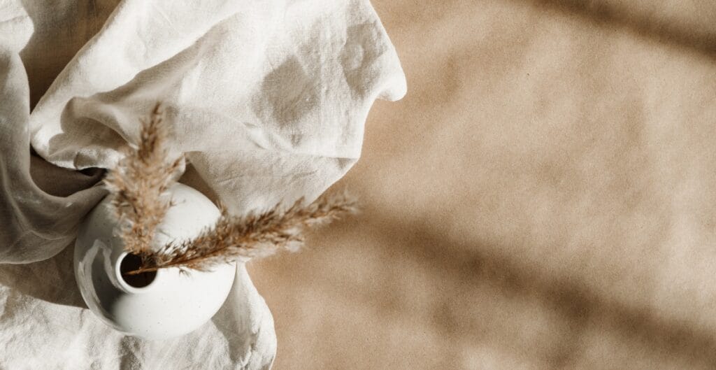 A white ceramic vase holding a few dried pampas grass stems, placed on a soft beige and cream fabric background.
