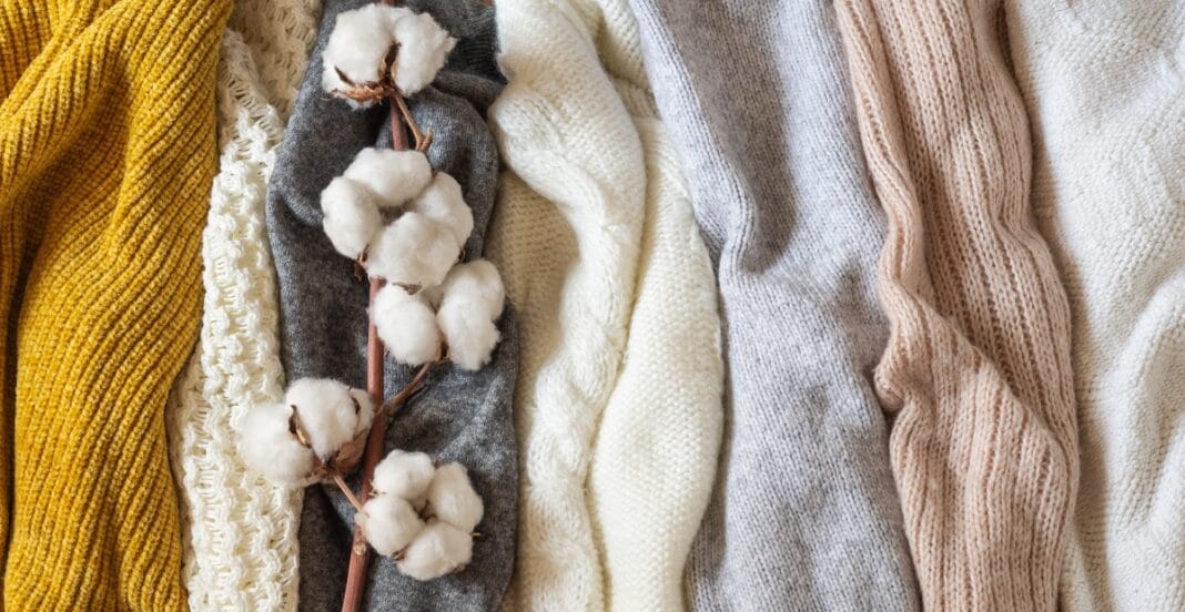 A close-up view of various knitted and woven fabrics in shades of mustard yellow, white, gray, and blush pink, with a cotton branch placed on top.